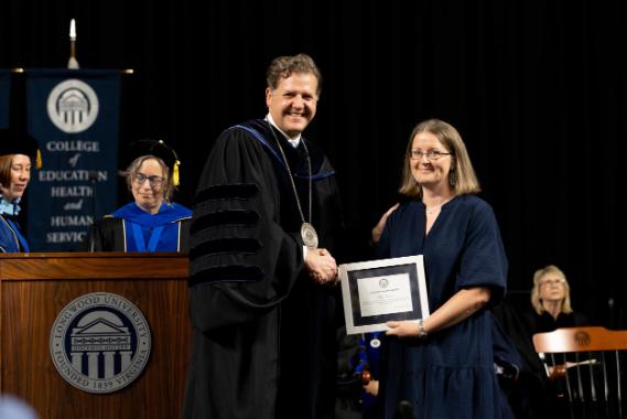 Mary - Two individuals shake hands on stage during a formal ceremony.   One wears academic regalia with a medallion; the other holds a framed certificate.   Banners with “COLLEGE” and column emblems are visible in the background.   Both are smiling, marking a moment of recognition or achievement.