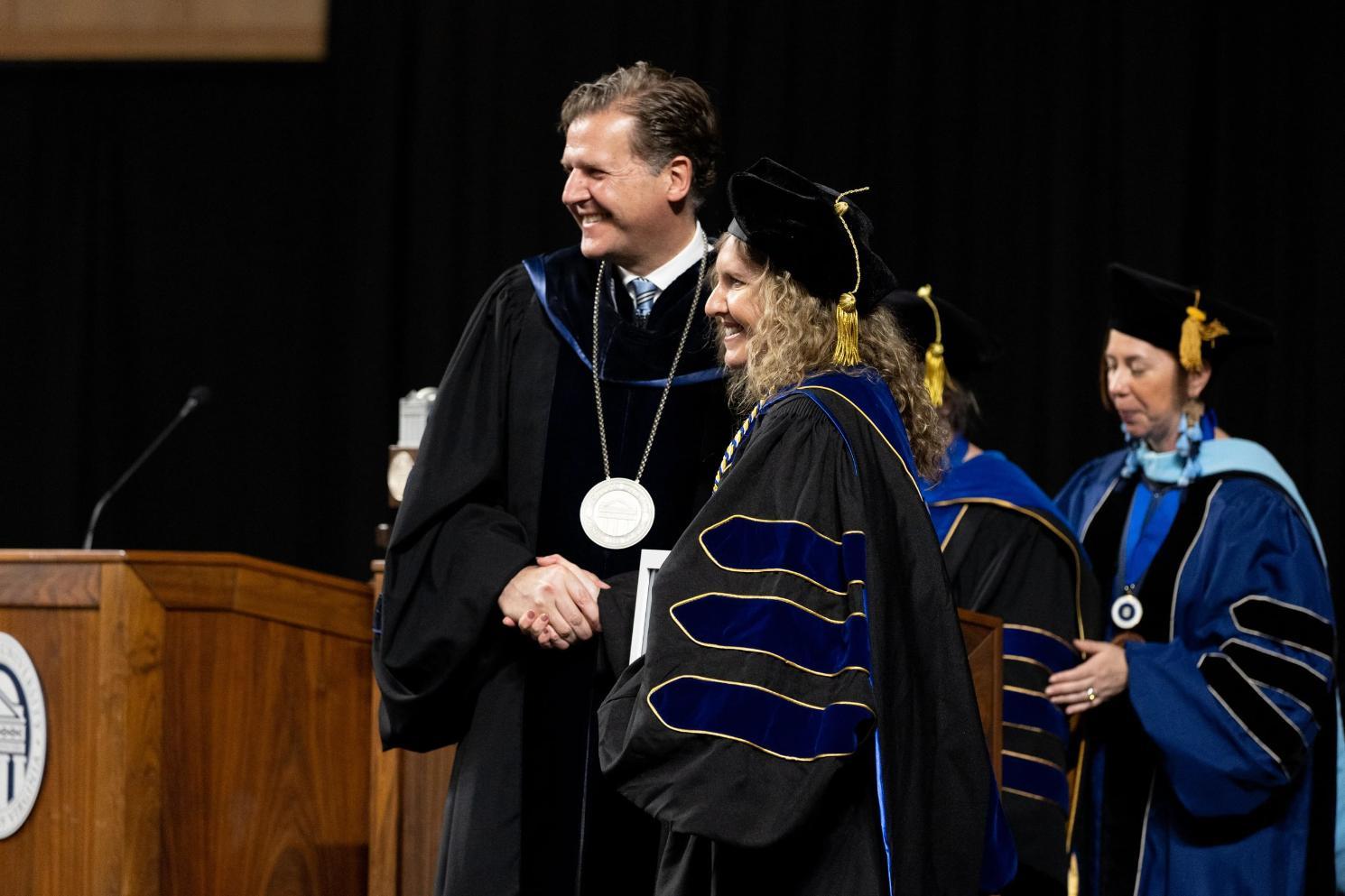 Awards - Two individuals shake hands on stage during a formal ceremony.   One wears academic regalia with a medallion; the other holds a framed certificate.   Banners with “COLLEGE” and column emblems are visible in the background.   Both are smiling, marking a moment of recognition or achievement.