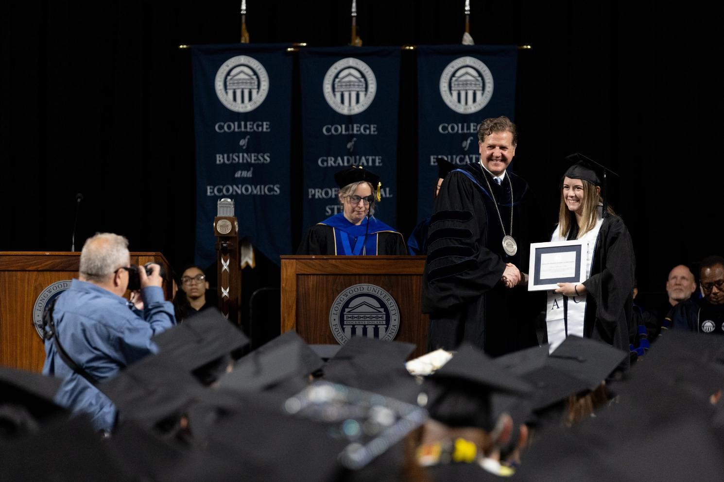 Katherine Keesee - Two individuals shake hands on stage during a formal ceremony.   One wears academic regalia with a medallion; the other holds a framed certificate.   Banners with “COLLEGE” and column emblems are visible in the background.   Both are smiling, marking a moment of recognition or achievement.
