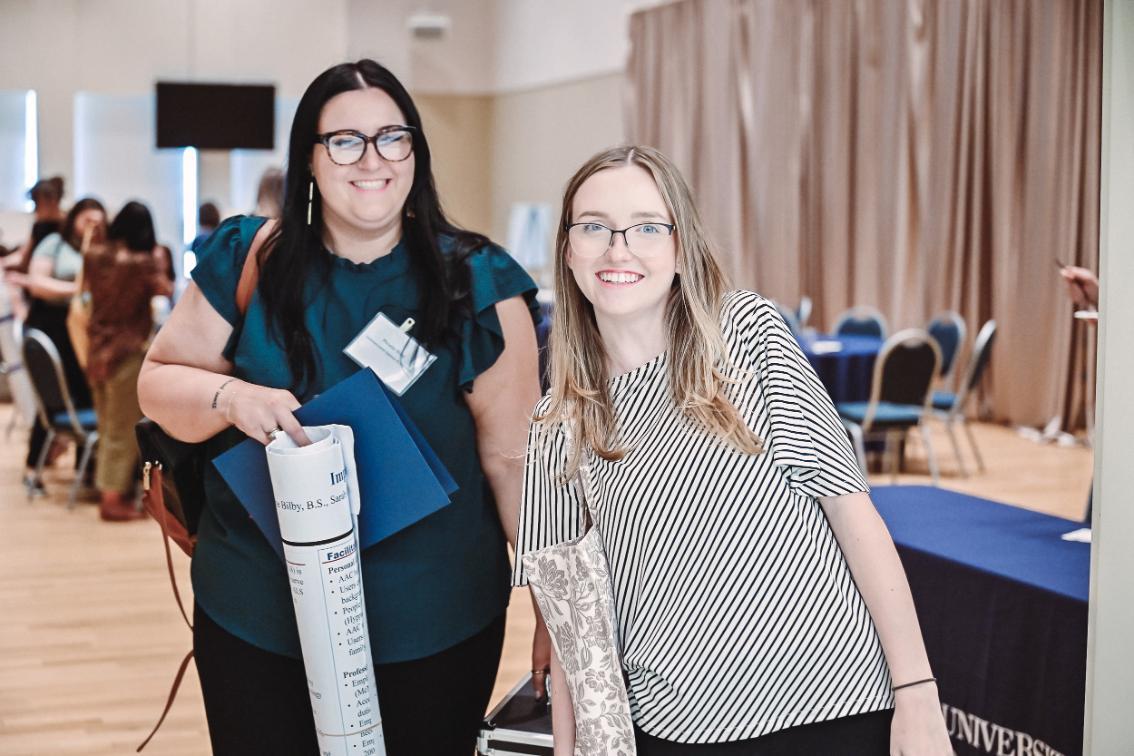 Posters - Two individuals stand indoors holding rolled-up posters and folders.   One wears a dark green top; the other wears a striped top.   The background includes people, chairs, and tables at an event.   The setting suggests a conference or presentation environment.