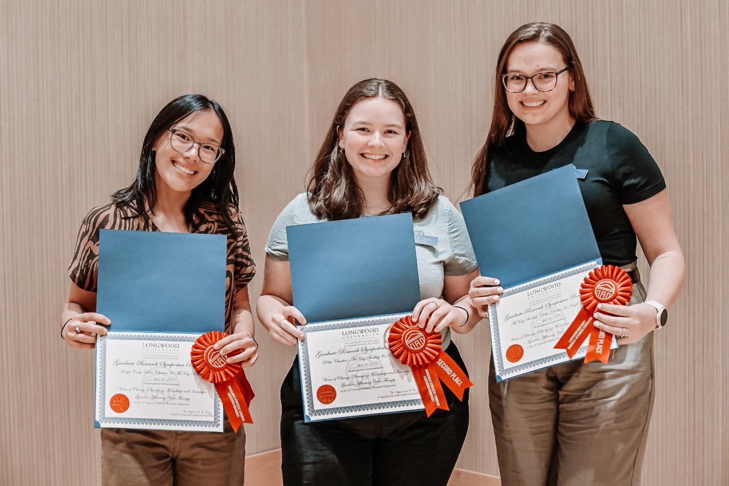 Group of 3 winners - Three individuals stand side by side holding certificates with red “HONORS” ribbons.   The certificates are from Longwood University, recognizing achievement.   They are smiling in an indoor award or presentation setting.   The background is plain, suggesting a formal ceremony space.