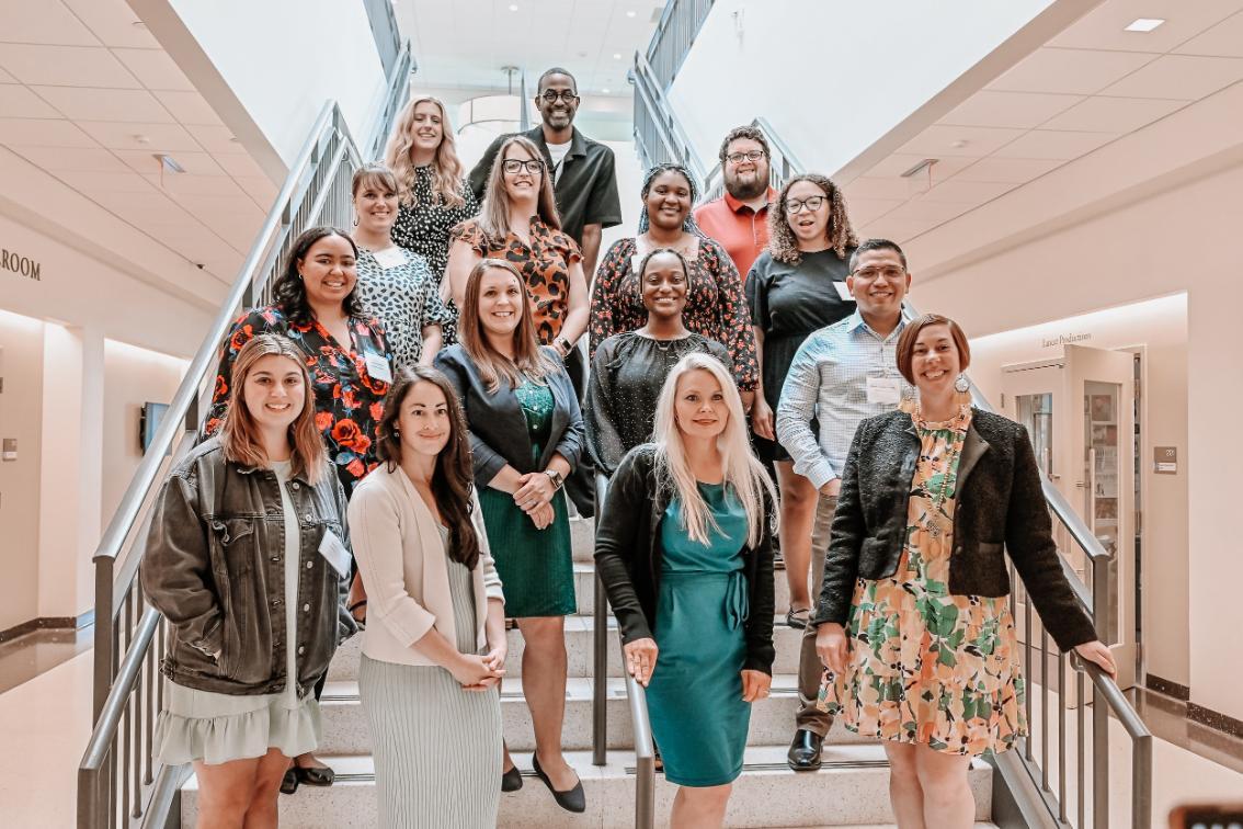EDLD Program - Fifteen people stand on an indoor staircase in three organized rows.   They wear a mix of casual and business attire in a professional setting.   The background includes doors and signage, suggesting an institutional space.   The photo appears to document a team or group event.