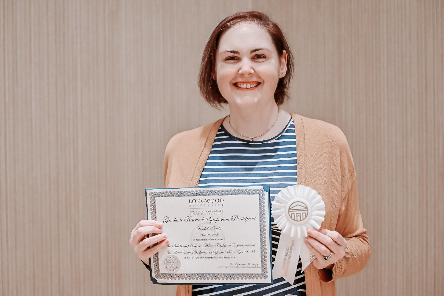 Rachel Toombs - A person stands holding a certificate and a white ribbon labeled “GRS.”   The certificate recognizes research on sleep deprivation and memory retention.   It was awarded at the Longwood University Graduate Research Symposium.   The individual is smiling against a plain indoor background.
