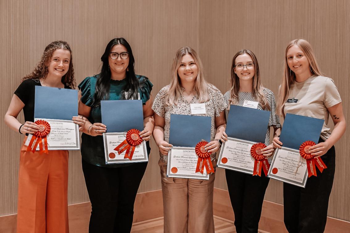 Group of 5 winners - Five individuals stand side by side holding certificates with red ribbons.   They are dressed in casual to semi-formal attire and smiling.   The setting appears to be an indoor award or recognition event.   The background is a plain wall, suggesting a formal presentation space.