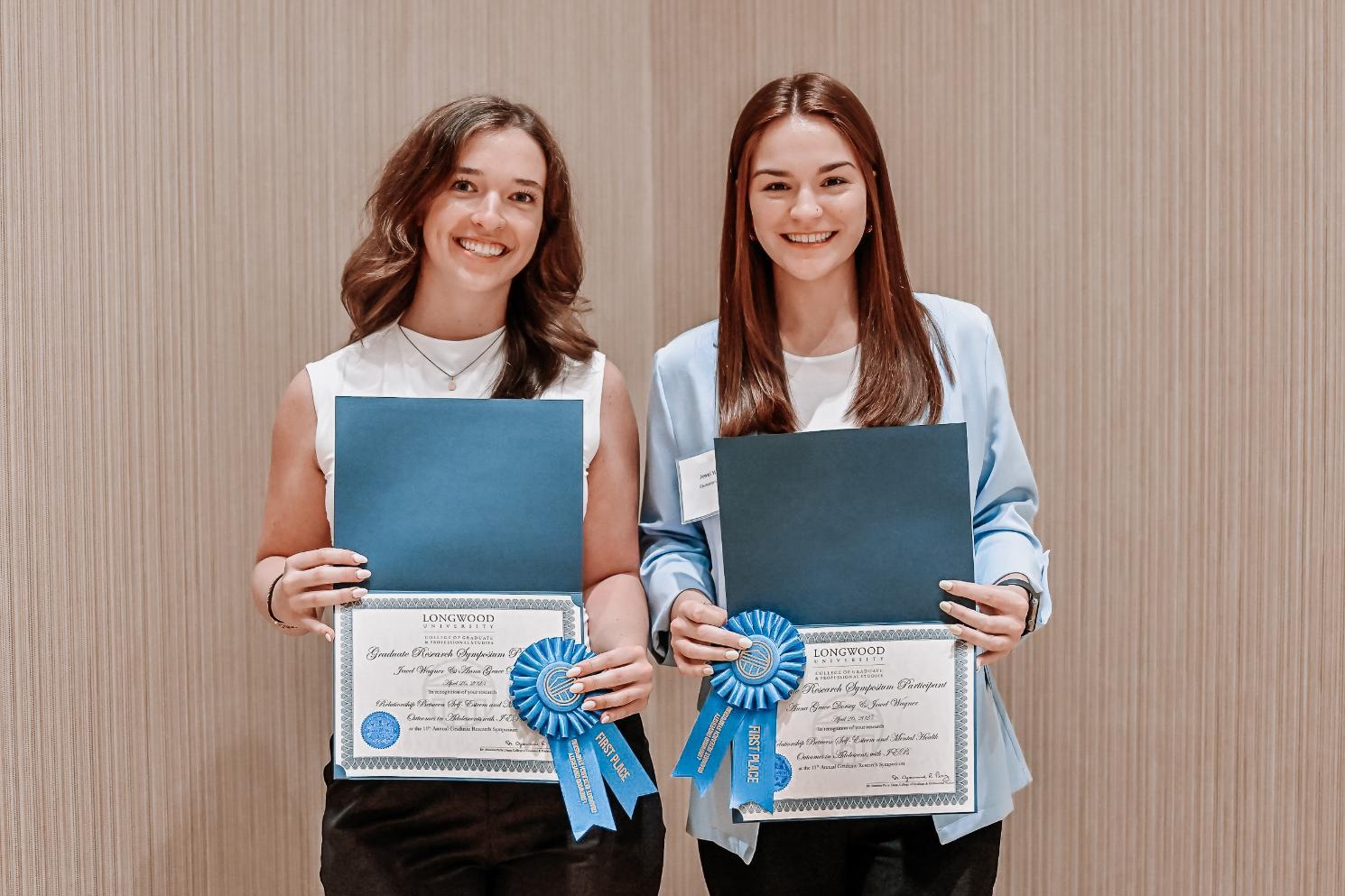 Jewel and Anna Winners - Two individuals stand side by side holding Longwood University certificates.   Each certificate has a blue ribbon labeled “FIRST PLACE” attached.   They are smiling in a celebratory award setting.   The scene highlights academic recognition and achievement.