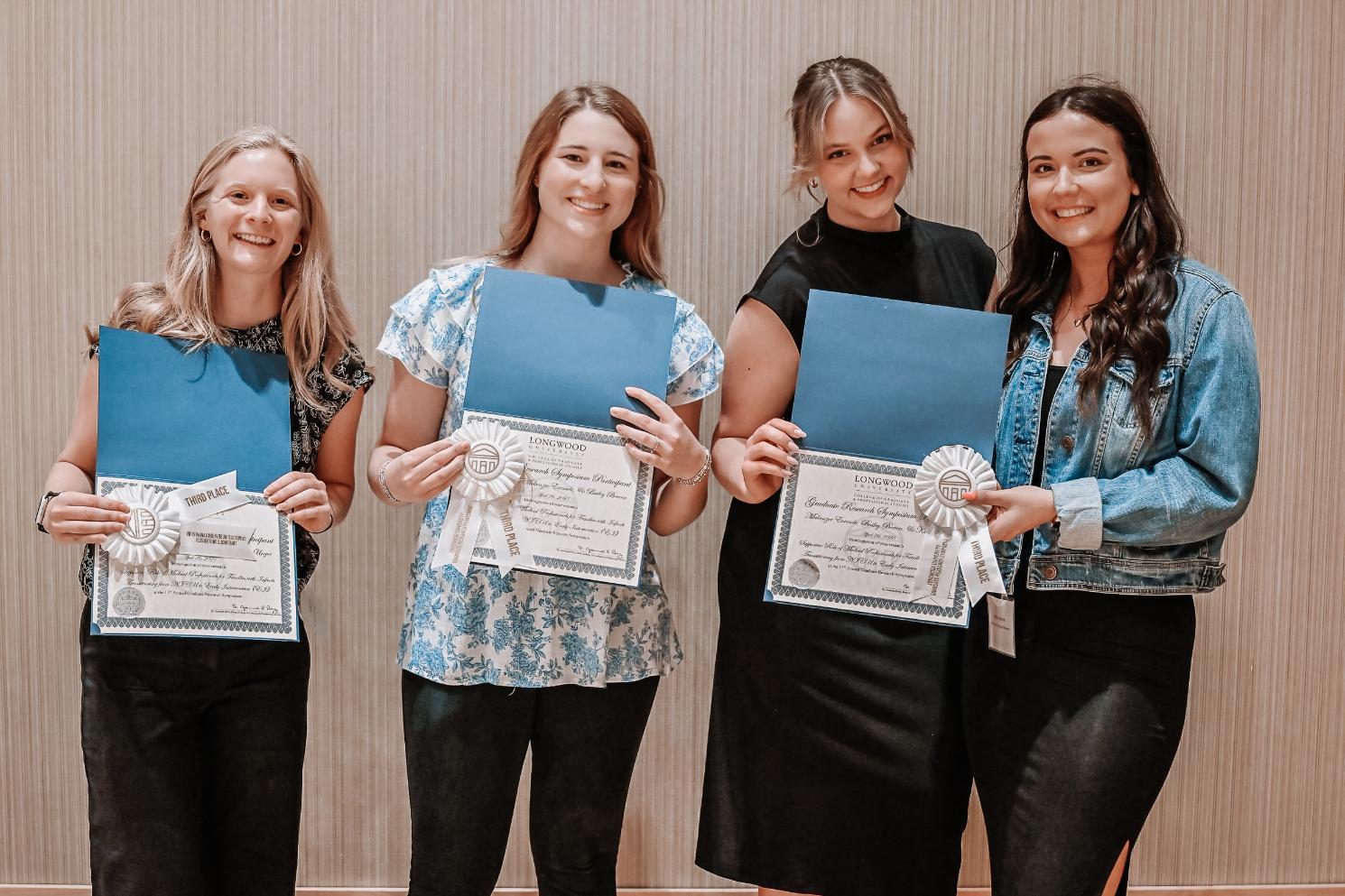 Kaley's Group Winners - Four individuals stand side by side holding certificates and blue folders.   Each certificate has a white ribbon attached, indicating recognition.   The setting appears to be an indoor award or achievement event.   The background features vertical lines, suggesting a formal venue.