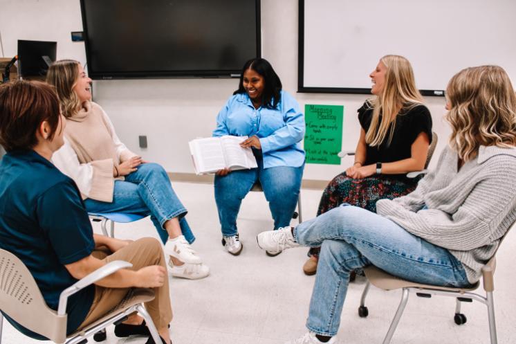 Workshop - Five people sit in a circle in a classroom, engaged in discussion.   One person holds an open book, appearing to read or present.   A large screen and green poster are visible in the background.   The room has white walls and light flooring, creating a bright setting.