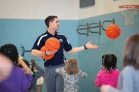 Health & Physical Education - A person stands in a gym holding multiple basketballs, surrounded by children.   The group appears to be participating in a recreational or PE activity.   A basketball hoop is visible in the background against blue and white walls.   The scene captures active engagement in a gymnasium setting.