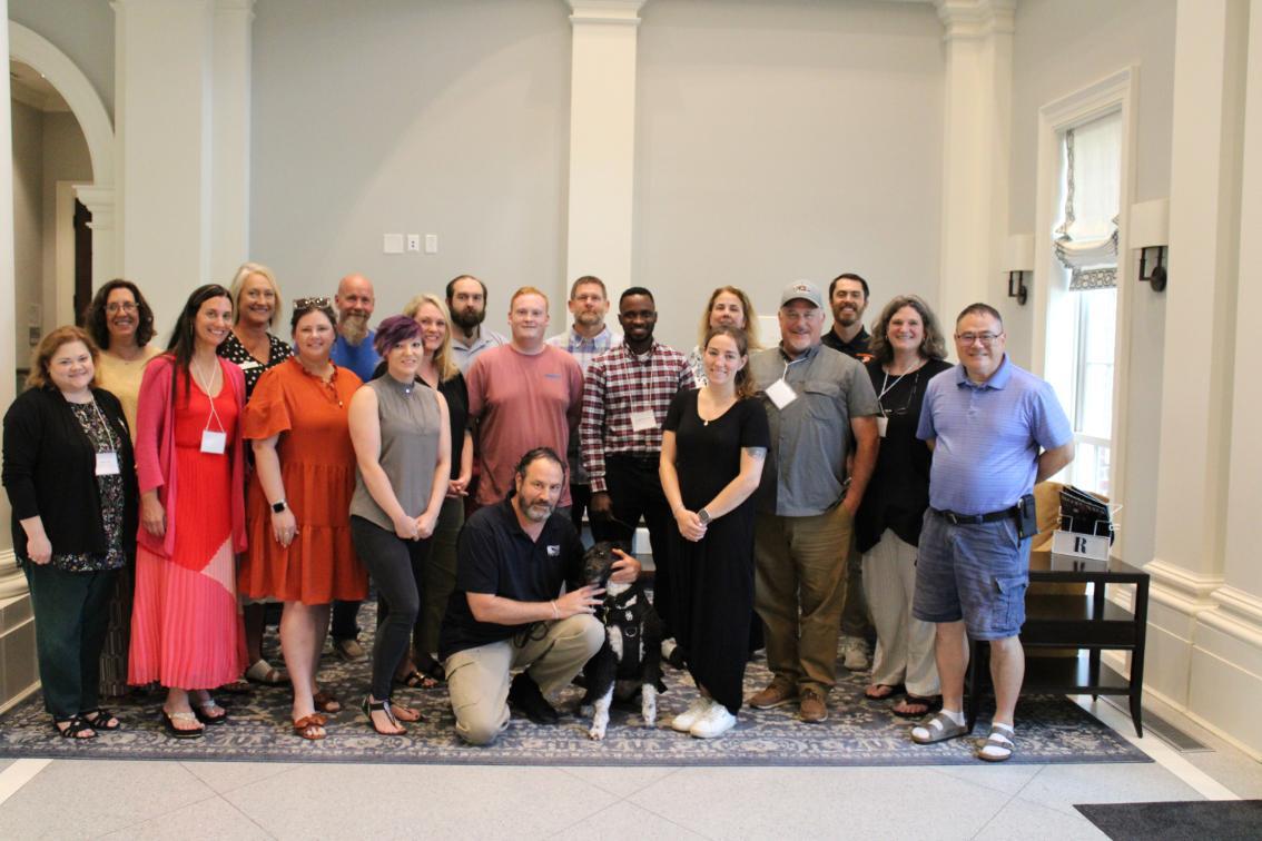 CTE Institute - Nineteen people pose indoors in two rows, with one seated holding a dog.   The setting features formal architectural details like columns and molding.   Attire ranges from casual to professional, suggesting a group or team event.   The arrangement includes standing, sitting, and kneeling individuals.