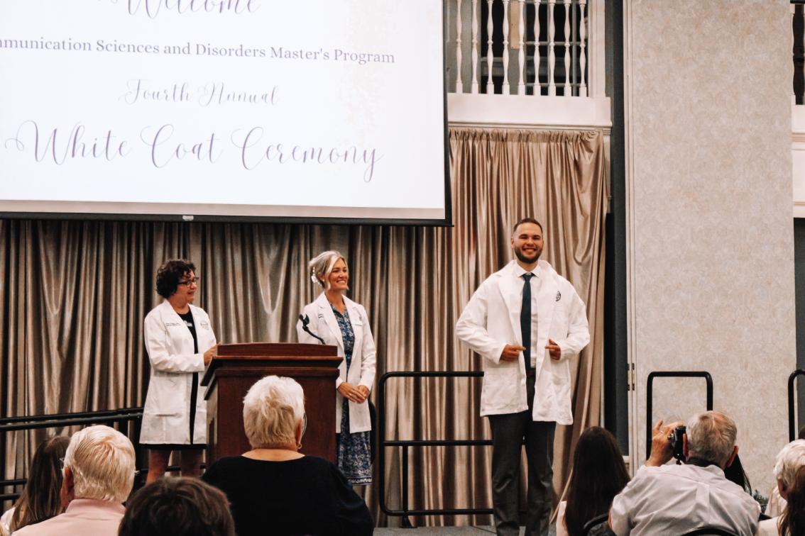 - Three individuals in white coats stand on a stage during a ceremony; one is putting on a white coat while the others stand near a podium, with a screen above reading “Welcome Communication Sciences and Disorders Master's Program Fourth Annual White Coat Ceremony” and an audience seated in front.