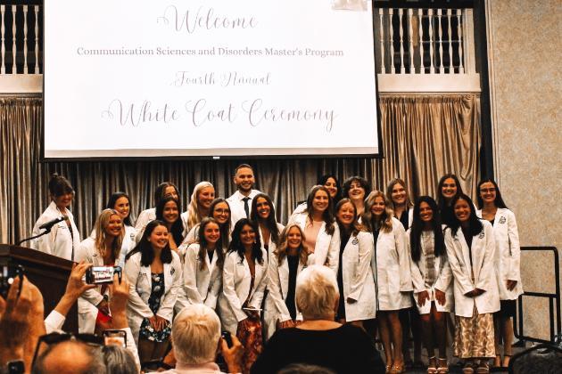 - A group of individuals in white coats stands on a stage beneath a screen that reads “Welcome Communication Sciences and Disorders Master's Program Fourth Annual White Coat Ceremony,” while audience members in the foreground watch and take photos.