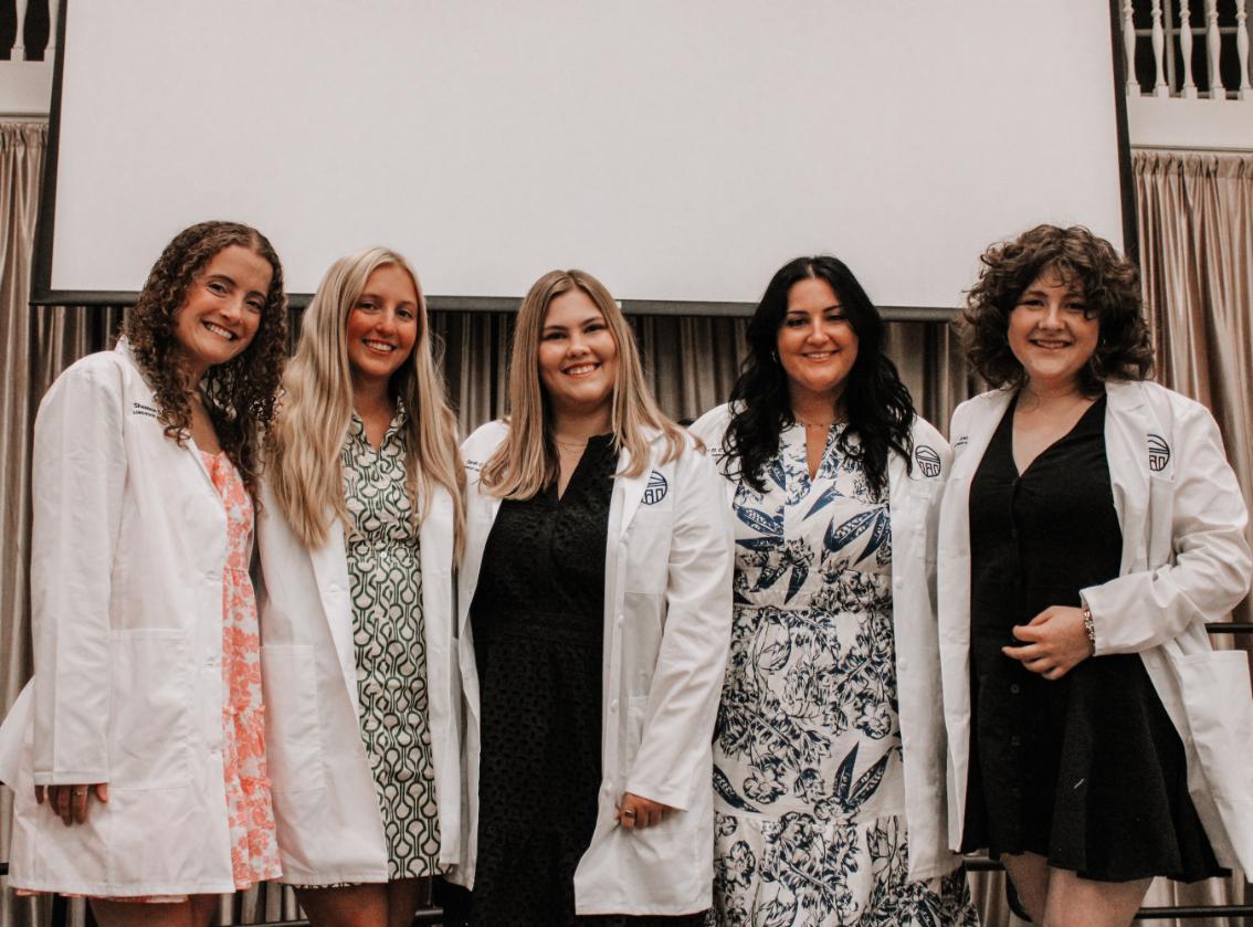 - Five individuals stand side by side wearing white lab coats over dresses, smiling in a formal setting with a large screen and curtains in the background.