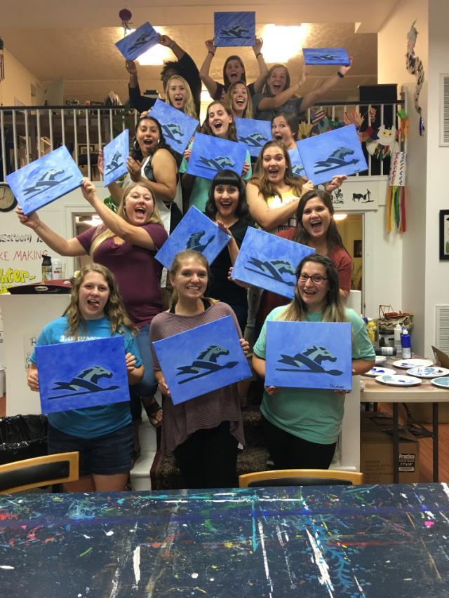 GSA Paint Night - A group of people pose indoors holding abstract blue and black paintings.   They are arranged in rows, with some standing on chairs or steps.   The background includes art supplies and decorations, suggesting a studio.   The scene reflects a shared creative activity or group art session.