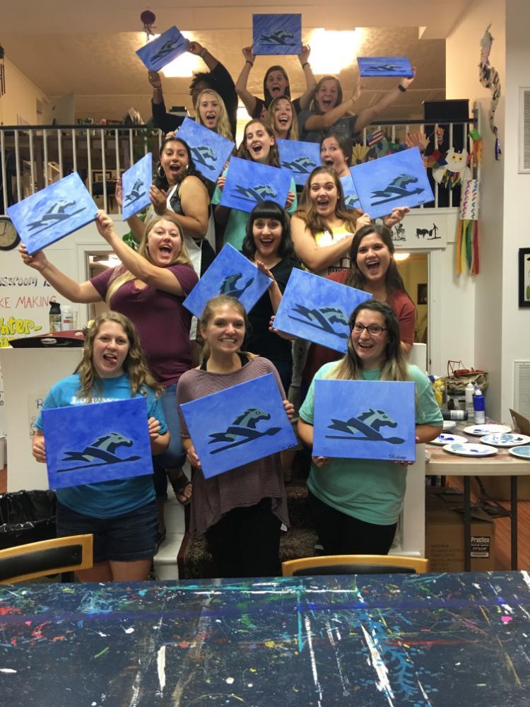 GSA Paint Night - A group of people pose indoors holding abstract blue and black paintings.   They are arranged in rows, with some standing on chairs or steps.   The background includes art supplies and decorations, suggesting a studio.   The scene reflects a shared creative activity or group art session.