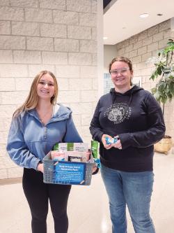Self-Care Station - Two people stand indoors in front of a brick wall during an appreciation event.   One holds a blue basket labeled “Happy Graduate Student Appreciation Week!”   The basket contains snacks and drinks; the other holds items in both hands.   Both appear to be celebrating graduate student recognition.