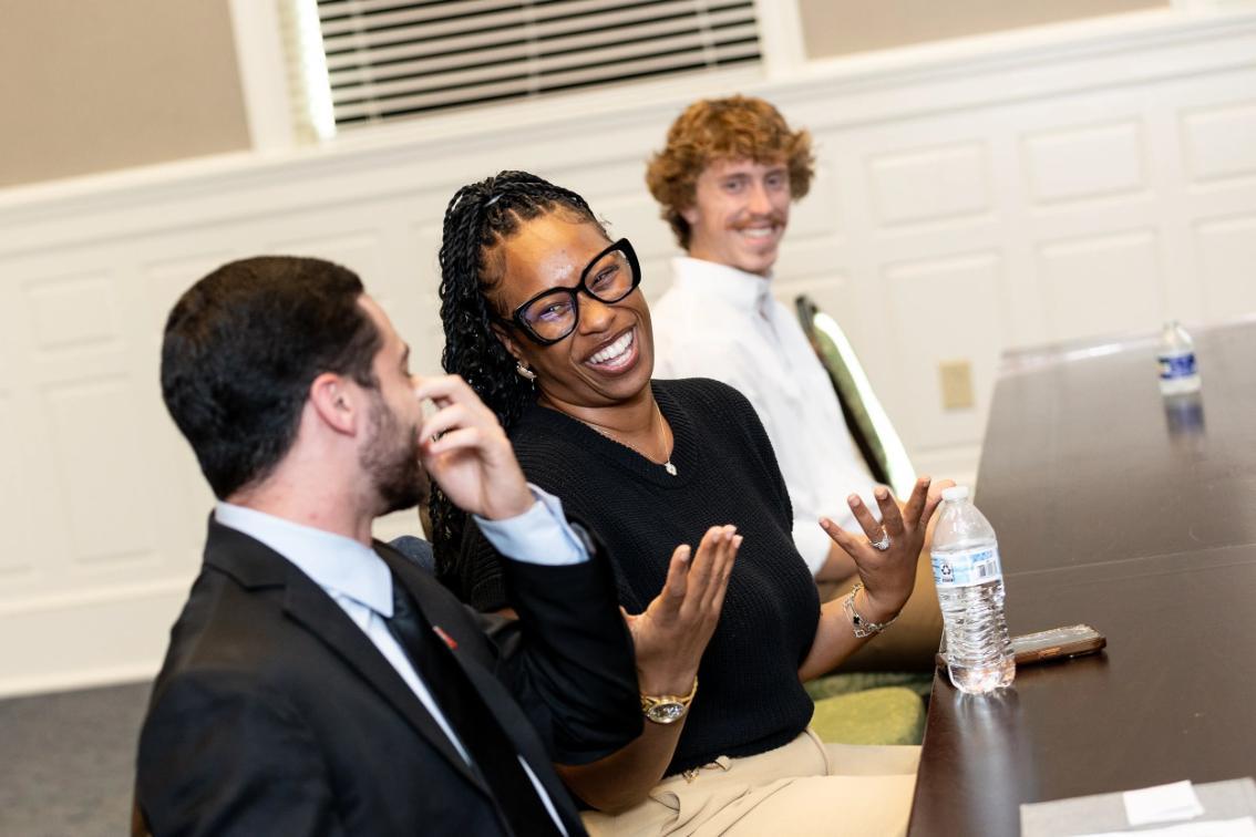 - Three people sit at a conference table in conversation; the person in the middle, wearing glasses and a black top, gestures while smiling, with a water bottle and smartphone on the table.