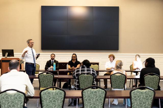 - A group of people sits at tables in a conference room facing a speaker who stands at the front gesturing, with a large blank screen behind them, suggesting a formal meeting or presentation setting.