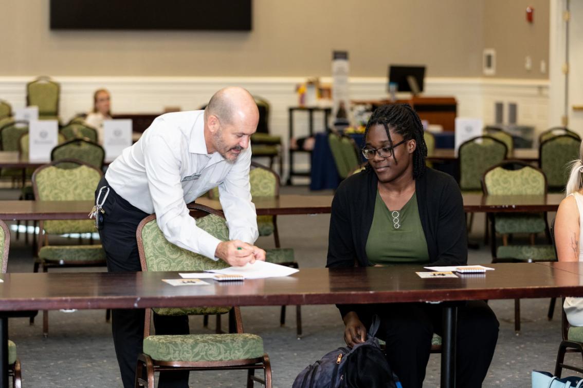 - A person in a white shirt leans over a table pointing at a paper while speaking to a seated individual with braided hair, glasses, a green top, and black cardigan, in a conference room with empty chairs and tables in the background.