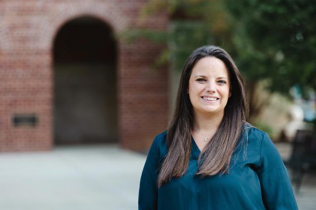 - A person with long straight hair wearing a dark green top stands outdoors smiling in front of a brick building with an arched entrance, with greenery and blurred background elements.