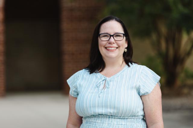 - A person with shoulder-length dark hair and glasses wearing a light blue short-sleeved striped blouse stands outdoors smiling, with brick walls and greenery blurred in the background.
