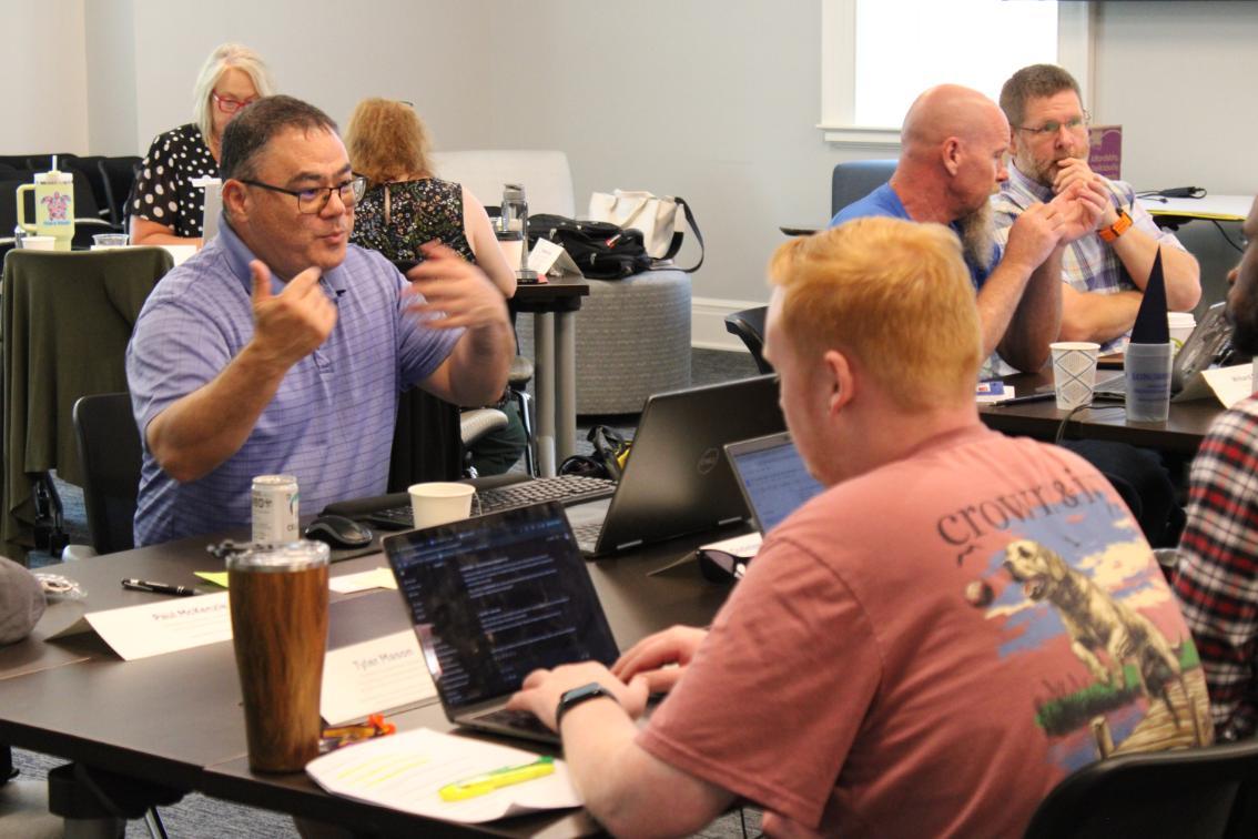 Paul McKenzie - A group of people sit around tables in a classroom or meeting room; two individuals in the foreground are focused, one gesturing while speaking and the other typing on a laptop, with others engaged in conversation or working in the background.