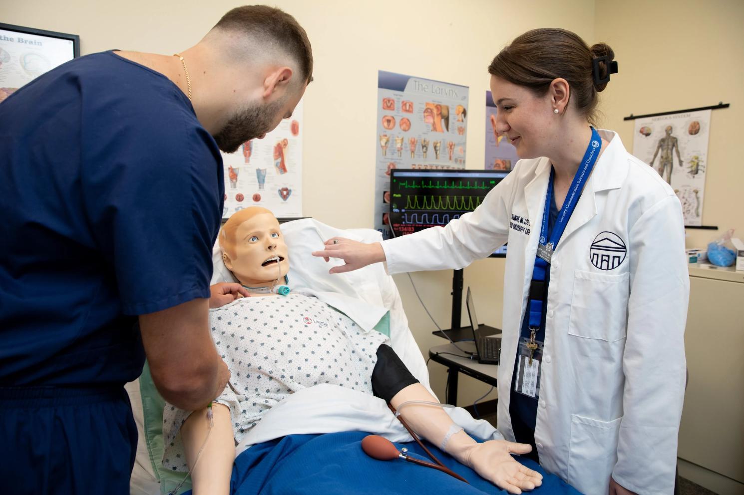 - Two individuals in a clinical setting work with a medical training mannequin on a hospital bed; one in blue scrubs adjusts the mannequin’s arm, while the other in a white lab coat touches its chest, with medical equipment and anatomical charts in the background.