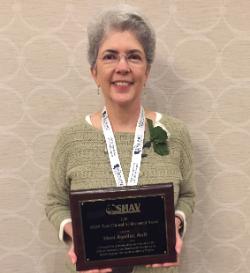 Dr. Mani Aguilar, State Clinical Achievement Award - A person holds a plaque for the 2019 SHAV State Clinical Achievement Award.   It honors Mani Aguilar for contributions to communication sciences.   They wear a SHAV lanyard and a green leaf pin on a sweater.   The background shows a patterned wall in an indoor setting.