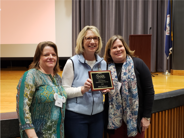 Judy Deichman receiving the VAASL James Regional Librarian of the Year Award - Three people stand on a stage with one holding a recognition plaque.   The plaque honors Judy Dickinson as VAASL School Librarian of the Year 2019.   The setting includes a wooden floor, podium, and curtain backdrop.   All are dressed professionally in an indoor award presentation setting.