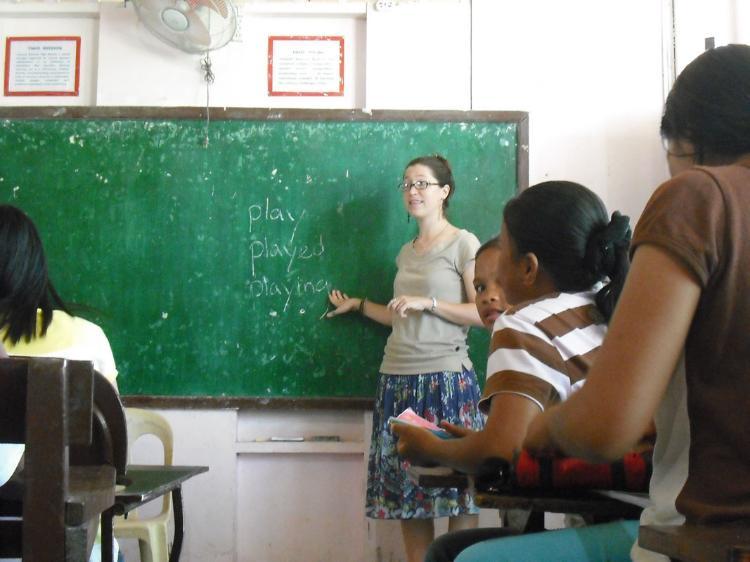 Jeannie Pfautz teaching in the Philippines. - A teacher stands at a chalkboard pointing to the words “play,” “played,” and “playing.”   Students sit at desks facing the teacher, engaged in the lesson.   The classroom includes a wall-mounted fan and framed posters.   The setting reflects a structured learning environment.