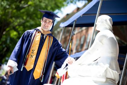 An undergraduate places his had on Joney on the Stoney as he leaves the Commencement stage. - A graduate in a navy cap and gown stands beside the statue of Joan of Arc, smiling in the sunlight. He wears gold and blue honor cords and a gold stole with Greek letters, touching the statue’s base in a celebratory gesture. The scene captures a Longwood University tradition during commencement on Brock C