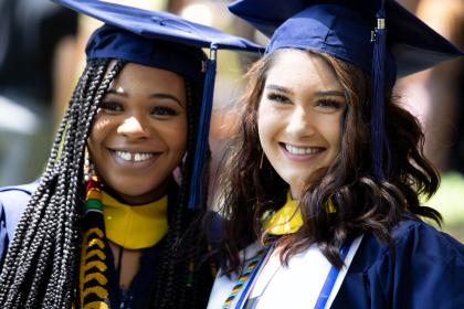 A close up of two graduates wearing caps and smiling for the camera.  - Two graduates in navy caps and gowns smile together in the sunlight. They wear yellow hoods and stoles with colorful cords, standing close in celebration. The soft focus background and natural light highlight their joyful expressions on graduation day.