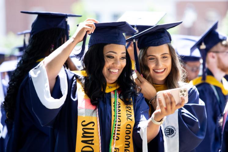 two students smile and take a selfie - Two graduates in navy blue caps and gowns smile as they take a selfie during the commencement ceremony. One adjusts her cap while the other holds the phone, both wearing gold stoles and honor cords. Sunlight brightens their faces as rows of fellow graduates stand behind them in celebration.