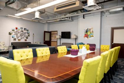 Photo of interior of Stevens Hall; Thinker, Maker, Creator Space - A modern lounge and meeting space with bright yellow chairs around a wooden table. Purple baskets and tissues sit neatly on the tabletop. Comfortable sofas, artwork, and a TV line the back wall under exposed ductwork. The room is well-lit with natural and overhead light, creating a welcoming atmosphere.
