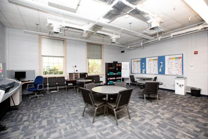 Photo of interior of Stevens Hall; Thinker, Maker, Creator Space - A spacious classroom or study area with round tables and office chairs. Large windows let in natural light along one wall. A bulletin board, whiteboard, and computer workstation line the perimeter. The room has gray carpet, exposed ductwork, and a clean, modern design.