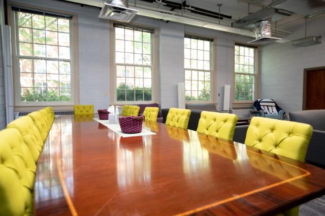 Honors Space - Stevens Hall - A bright meeting room with a long polished wooden table surrounded by yellow upholstered chairs. Large windows line the wall, filling the space with natural light. Purple woven baskets and tissues sit neatly on the table’s center runner. Exposed ceiling ducts and modern lighting add a contemporary touch to the room.