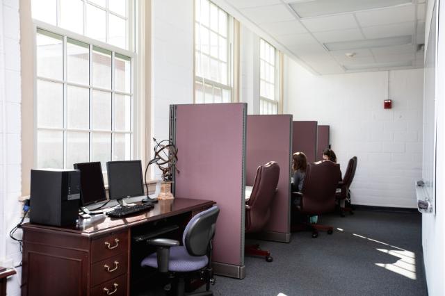 Honors Space - Stevens Hall - A quiet study area with tall windows letting in natural light. Several computer workstations and partitioned cubicles line the room. Students sit in brown leather chairs focused on their work. The atmosphere is calm and studious, with clean white walls and simple décor.