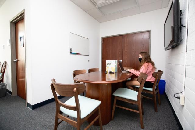 Honors Space - Stevens Hall - A student wearing a mask works on a laptop at a wooden conference table. The small meeting room has several chairs, white walls, and a mounted TV screen. A whiteboard hangs on the wall beside a closed wooden door. The space feels quiet and organized, suitable for study or discussion.