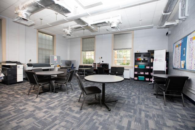 Honors Space - Stevens Hall - A bright, spacious office with round tables and black chairs arranged for group work. Printers and computers line the walls beneath large windows letting in natural light. A whiteboard and bulletin board display notes and schedules. The room has a clean, organized look with exposed ductwork and modern furnishings.