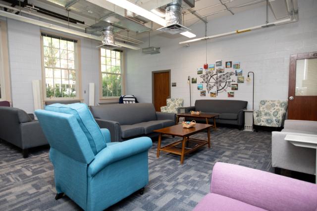 Honors Space - Stevens Hall - A bright, modern lounge space with colorful armchairs and gray sofas arranged around wooden coffee tables. Large windows let in natural light, illuminating artwork and photos on the far wall. Exposed ductwork and light fixtures give the room an industrial yet comfortable feel. Snack baskets and soft furnishings create a welcoming, relaxed atmosphere.