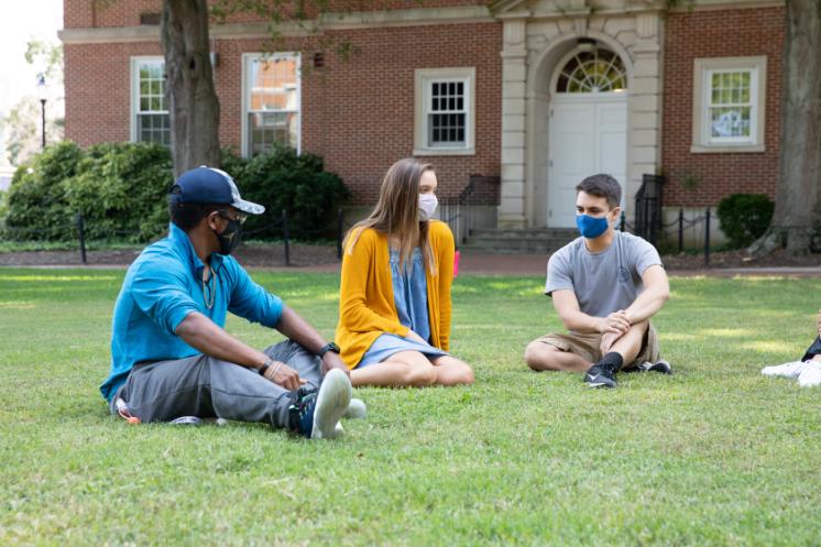 students sit outside stevens hall - Three students sit on the grass in front of a red-brick campus building, spaced apart and wearing face masks. They appear to be engaged in conversation, enjoying a casual outdoor class or meeting. Sunlight filters through the trees, casting soft shadows on the lawn. The setting feels calm and studious, reflecting a socially distanced campus atmosphere.