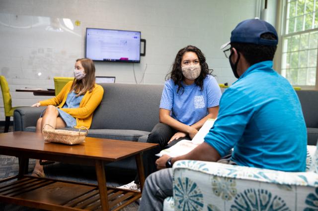Honors Space - Stevens Hall - Three students sit spaced apart in a bright lounge area, wearing masks and talking. One student in a blue Cormier Honors College T-shirt faces two others seated on sofas. A TV screen and whiteboard are visible on the wall behind them. Natural light fills the room through large windows, creating a relaxed, collaborative atmosphere.