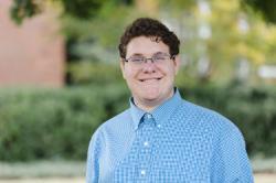 Portrait of a young man with short curly brown hair and glasses, smiling while wearing a blue checkered button-up shirt, standing outdoors with green trees and blurred foliage in the background.