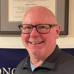 -Person smiling indoors in front of framed certificates on the wall. They are wearing glasses and a dark zip-up pullover with a visible Longwood University logo. The lighting is warm, highlighting their face clearly. The background includes a navy banner and a diploma in a dark frame.