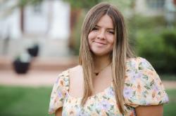 Cameron Lowery -Person smiling outdoors on a college campus. Wearing a floral dress with puff sleeves and a gold necklace. Straight light brown hair parted in the middle. Soft background with greenery and blurred campus buildings.