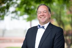 Drew Hudson -Man wearing a dark suit, white shirt, and patterned bow tie smiling outdoors. He stands in front of a softly blurred background of trees and greenery. Natural light highlights his face and professional appearance. The photo conveys a warm and approachable tone.
