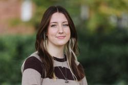 Portrait of a young woman with long brown hair and blonde highlights, wearing a brown and beige striped sweater and layered necklaces, smiling softly outdoors with a blurred background of greenery.