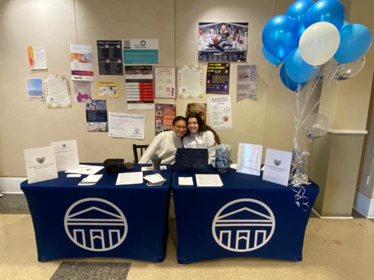 Student Philanthropy Council members Becca Chung '23 and Kensington McLoone '24 engage with Lancers on National Philanthropy Day. -Two people sit behind tables covered in blue cloths with the Longwood University logo, smiling at a National Philanthropy Day display. The tables feature informational signs, papers, pens, and a jar of candy, with blue and white balloons to the side. Colorful campus fl