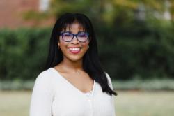 Portrait of a young woman smiling outdoors, wearing blue glasses, a cream-colored sweater, and a gold necklace with a cross pendant. The background is softly blurred with green foliage and a hint of sunlight filtering through the trees.