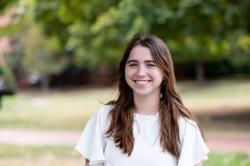 Portrait of a young woman smiling outdoors on a sunny day, wearing a white blouse with flutter sleeves. She stands in front of a softly blurred background of green trees and grass, creating a warm, natural campus setting.