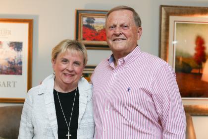 -An older couple stands together indoors, smiling warmly at the camera. The woman wears a white textured jacket over a black top with a cross necklace. The man wears a pink-and-white striped button-down shirt. Framed artwork with landscapes and bright colors hang on the wall behind them.